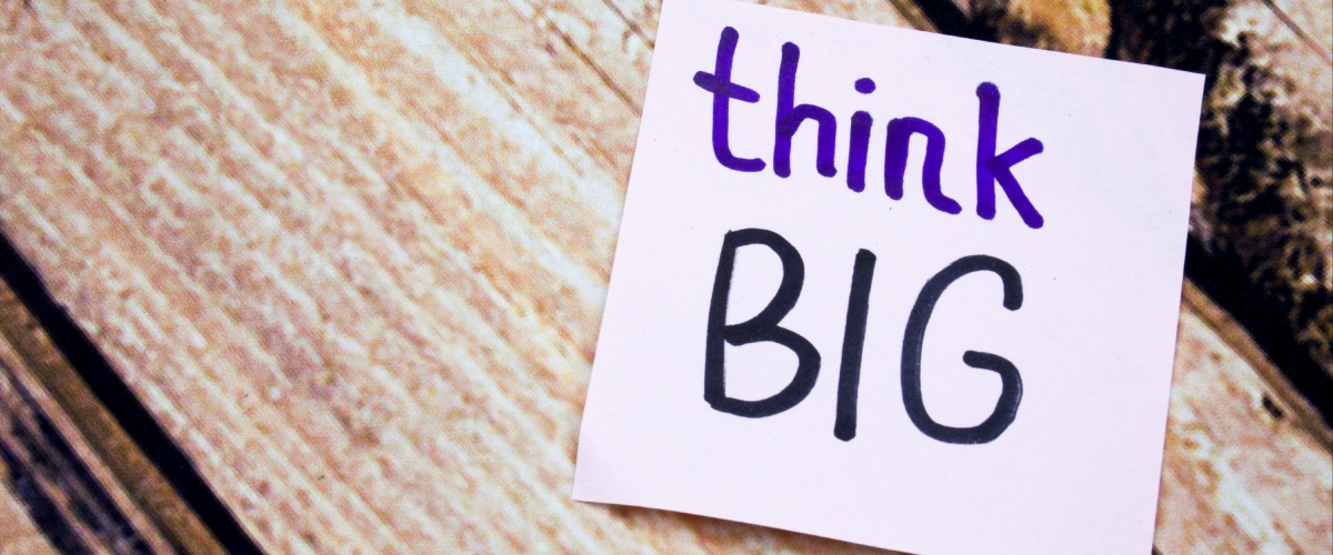 A purple note sits in front of a wooden background. The note's text says "think big."