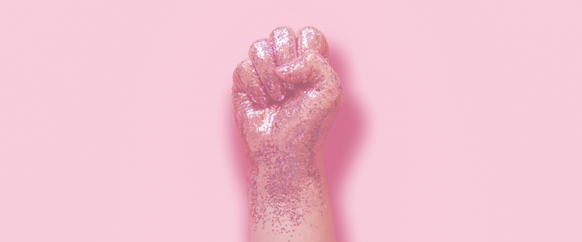 A pink, glittery fist is in front of a pale pink background. 