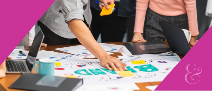 People are standing and working around a table. There are sticky-notes, computers, books and posters. A poster says "brand."