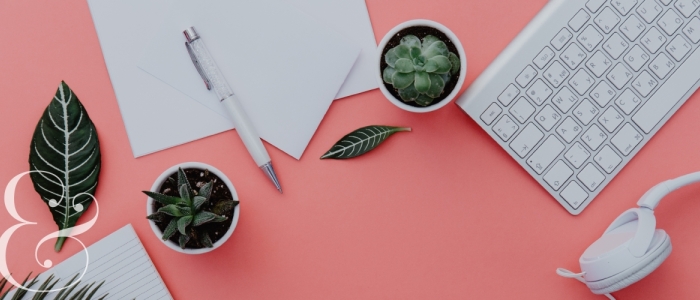 Dark green plants, office supplies, and a keyboard and headphones sit on top of a pink desk. A white ampersand is in the bottom left corner. 